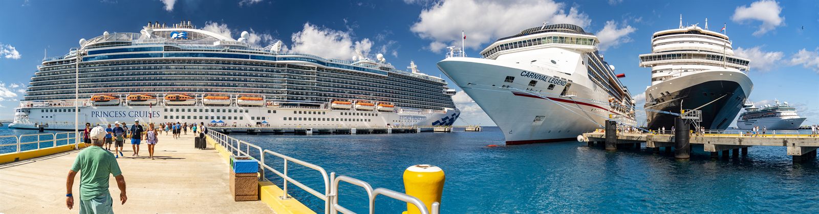 Passengers walking along a pier toward several docked cruise ships
